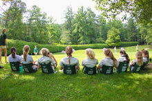 Load image into Gallery viewer, 9 girls sitting in dark green crazy creek chairs in a line on an archery field on a sunny day. 
