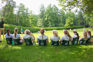 9 girls sitting in dark green crazy creek chairs in a line on an archery field on a sunny day. 