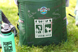 Dark green crazy creek chair, that has been signed with many names, sitting in grass with the square Merrie-Woode logo of a horse, its rider, and two girls on a dock on the back of the chair.