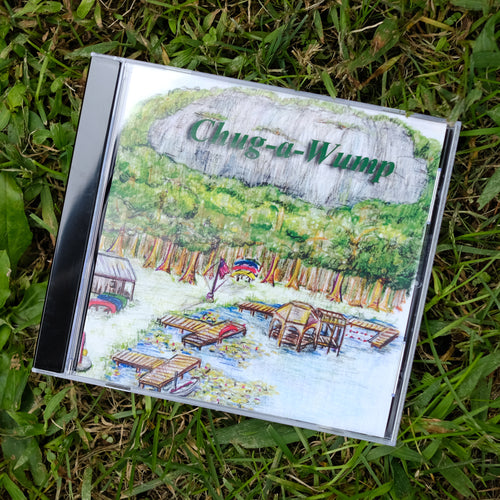 CD case sitting in green grass. CD cover shows a drawing of three docks on a lake with 8 canoes under the Old Bald mountain face, which is surrounded by light green trees.