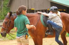Load image into Gallery viewer, Young, blonde girl wearing a black helmet,  jeans, a grey middie, and tall black boots is mounting a light brown horse. A young woman riding instructor, who is wearing a green polo shirt and khaki shorts, is standing next to the horse&#39;s face. 
