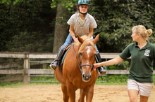 Load image into Gallery viewer, Young girl, who is wearing a green helmet, grey middle, blue jeans, and tall black boots, is riding a light brown horse. A blonde, woman riding counselor, who is wearing khaki shorts and a dark green polo, is walking next to the horse
