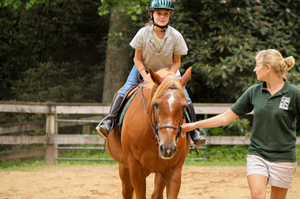 Young girl, who is wearing a green helmet, grey middle, blue jeans, and tall black boots, is riding a light brown horse. A blonde, woman riding counselor, who is wearing khaki shorts and a dark green polo, is walking next to the horse