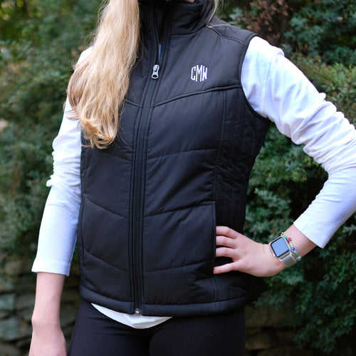 Blonde girl standing in front of a green bush, wearing a while long sleeve shirt with a black, puffer vest over that has the initials 