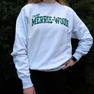 Blonde girl wearing a grey, crew neck sweatshirt that says "Camp Merrie-Wodoe" across the chest in green lettering. Girl is standing in front of dark green bushes.