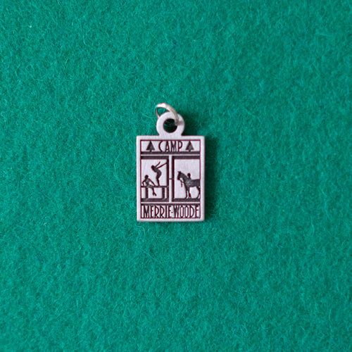 Small, grey charm, made of Pewter, that has the rectangle Merrie-Woode logo engraved into it. The logo shows a horse, its rider, and two girls on a dock. The charm is lying on a green background.