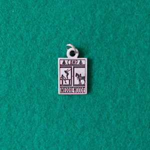 Small, grey charm, made of Pewter, that has the rectangle Merrie-Woode logo engraved into it. The logo shows a horse, its rider, and two girls on a dock. The charm is lying on a green background.