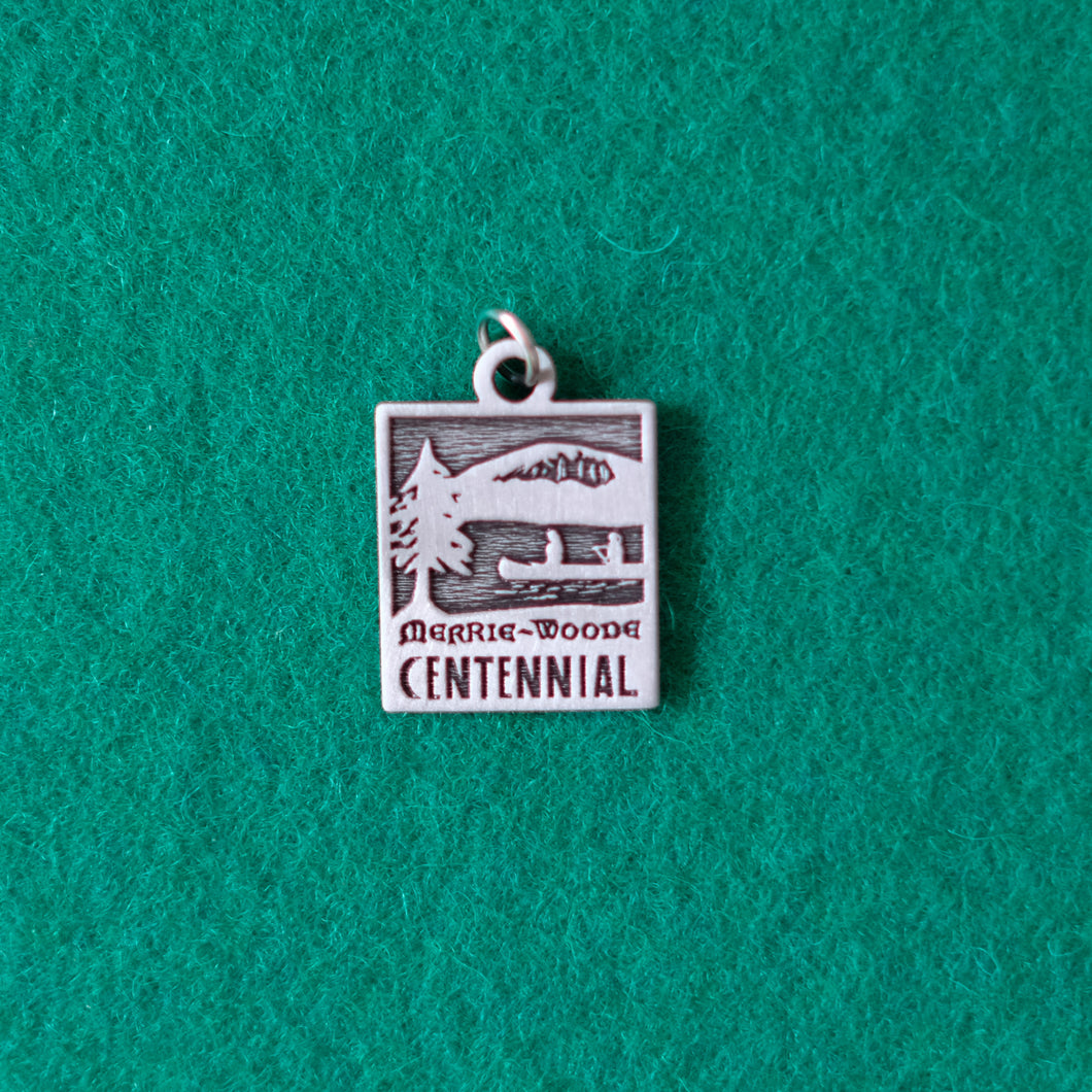 Small, pewter necklace charm with the Merrie-Woode Centennial logo, showing the Old Bald Mountain, two people paddling a canoe on lake Fairfield, and a tall tree. 