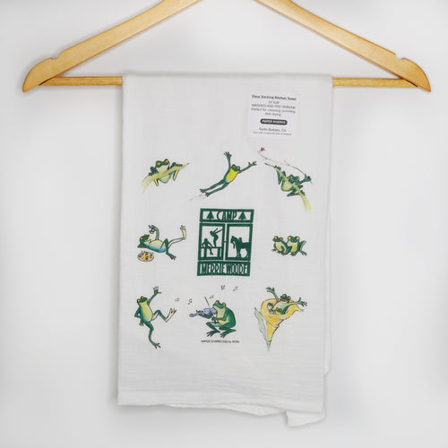 White kitchen towel with the green Merrie-Woode square logo in the middle, which shows a horse, its rider, and two girls on a dock. Around the logo are pictures of frogs jumping, playing the violin, eating, and dancing. 