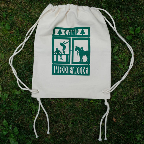 Tan canvas drawstring bag, with the Merrie-Woode rectangle logo, which shows a horse, its rider, and two girls on a dock. Canvas bag lying flat on green grass. 