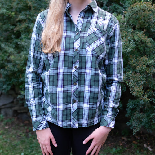 Blonde girl wearing a green and white long sleeve flannel with a white embroidered diamond Merrie-Woode logo on the upper right shoulder area. Girl in standing outside in front of green bushes. 