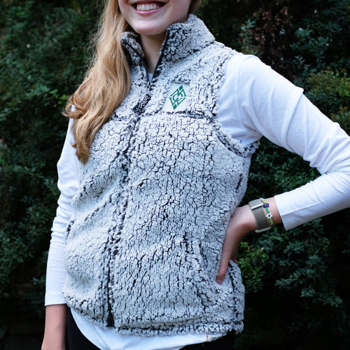 Blonde girl smiling wide, wearing a long sleeve white shirt, with a grey, fuzzy, Sherpa vest over. She is standing in front of green bushes.