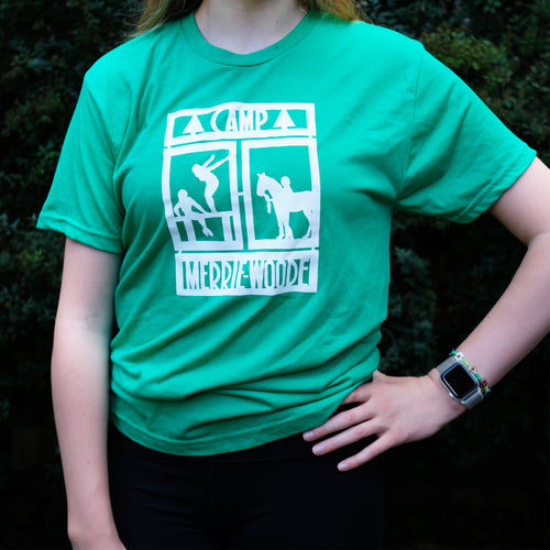 Blonde girls standing in front of dark green bushes wearing a green, short sleeve tee that has the rectangle Merrie-Woode logo, which shows a horse, its rider, and two girls on a dock.