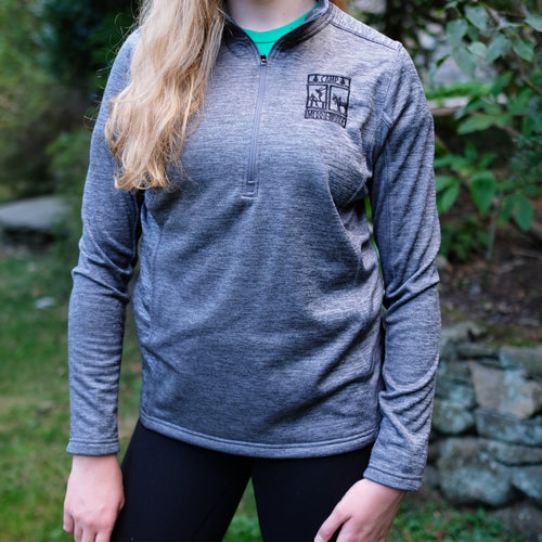Blonde girl wearing a grey 3/4 zip athletic pull over that has the rectangle Merrie-Woode logo on the top right, which has a horse, its rider, and two girls on a dock. The girl is standing in front of green trees.
