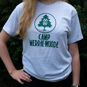 Blonde girl wearing a short sleeve grey tee that says "Camp Merrie-Woode" with the initials "CMW" in a green tree sketch at the top. Girl is standing in front of a green bush.