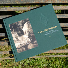 Load image into Gallery viewer, Green Camp Merrie-Woode Centennial coffee table book. Cover of book says &quot;Camp Merrie-Woode&quot; in gold above the phrase &quot;one hundred years of following the gleam.&quot; The right side cover of the book shows a waterfall and at the base there are canoes filled with Merrie-Woode campers. Book is sitting on a metal bench over light green grass. 
