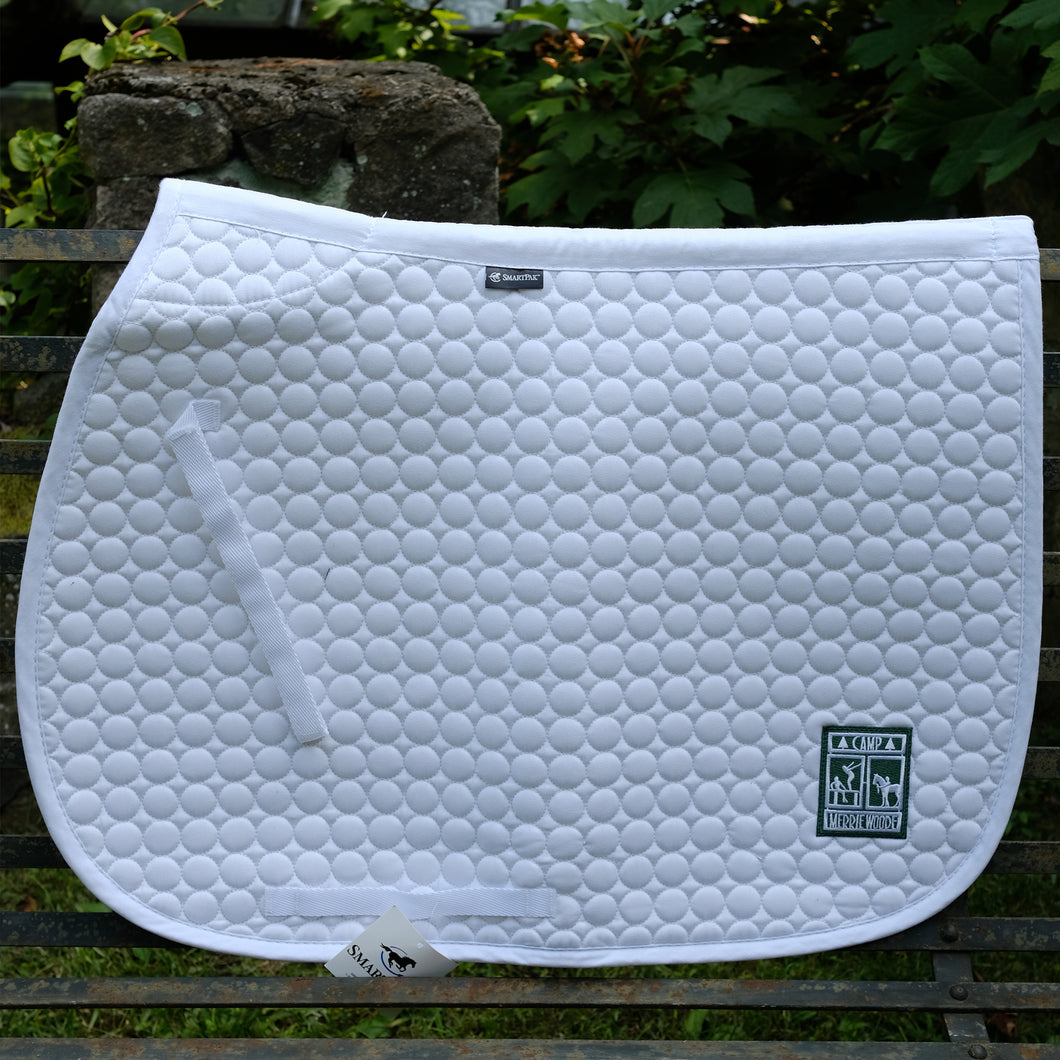 White saddle pad with the rectangle Merrie-Woode logo in the bottom right corner, that shows a horse, its rider, and two girls on a dock. Saddle pad is sitting on a metal bench in front of green leaves.
