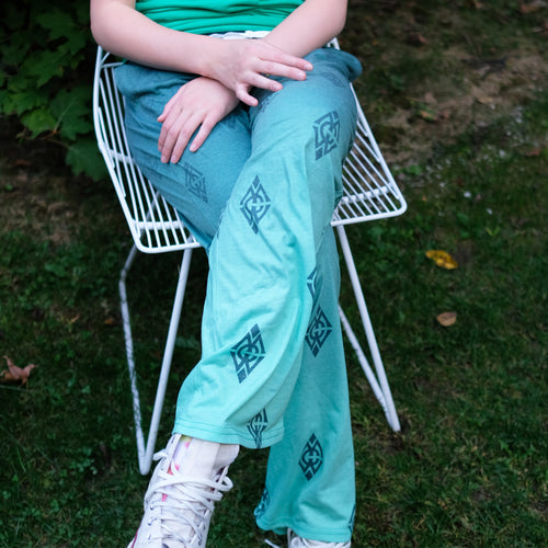 Picture showing lower half of a girl who is sitting in a white, wire chair, outside on green grass. Girl is wearing green ombre pajama pants that have the diamond Merrie-Woode logo throughout the fabric. She is wearing white converse shoes.