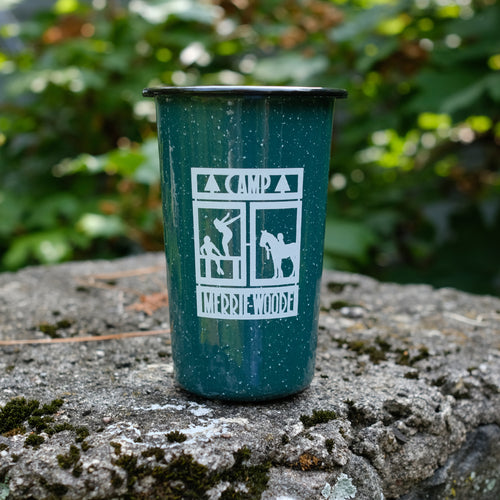 Green camping tin cup with the square Merrie-Woode logo, which shoes a horse, its rider, and two girls on a dock. Tin cup is sitting on a stone in front of green trees.