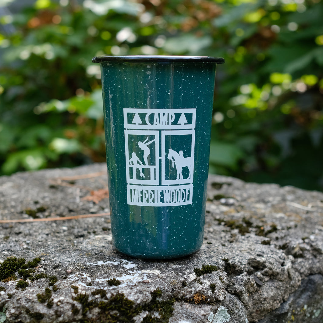 Green camping tin cup with the square Merrie-Woode logo, which shoes a horse, its rider, and two girls on a dock. Tin cup is sitting on a stone in front of green trees.