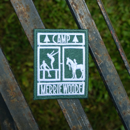 Patch of Camp Merrie-Woode rectangle logo (showing a horse, its rider, and two girls on a dock). Patch is on a wooden bench above green grass.
