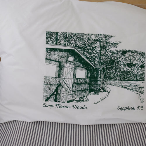 Picture of white pillow case that has a green picture on it showing the outside of 2 cabins, the Old Bald rock face, and the path in front of Lake Fairfield.