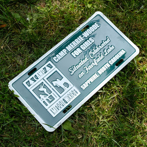 Green license plate that says: "Camp Merrie-Woode for Girls. Situated, Celebrated on Fairfield Lake. Sapphire, North Carolina." The right side of the plate has the white, square Merrie-Woode logo, which shows a horse, its rider, and two girls on a dock. 