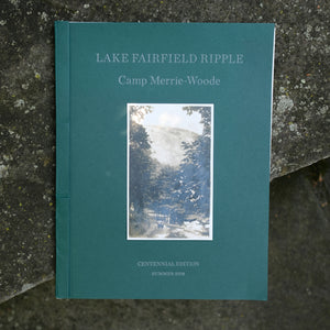 Dark green Merrie-Woode ripple book sitting on a stone. The ripple cover says "Lake Fairfield Ripple. Camp Merrie-Woode. Centennial Edition. Summer 2018." Cover also has a picture showing two campers walking down a tree-lined road with the Old Bald mountain above.