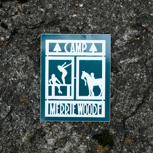 Green sticker with the rectangle Merrie-Woode logo, which has a horse, its rider, and two girls on a dock. Sticker is lying on rocky/gravel road.