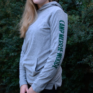 Blonde girl with her hair down is wearing a grey hoodie with the words "Camp Merrie-Woode" in green print going down the right arm sleeve. Girl is standing in front of green bushes. 