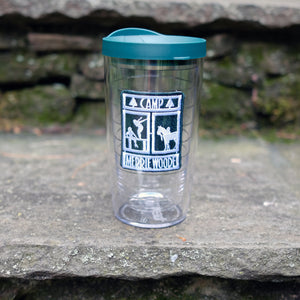 Clear Tervis tumbler with a green lid featuring the Camp Merrie-Woode rectangle logo, which has a horse, its rider, and two girls on a dock." Tumbler is sitting on a stone step.